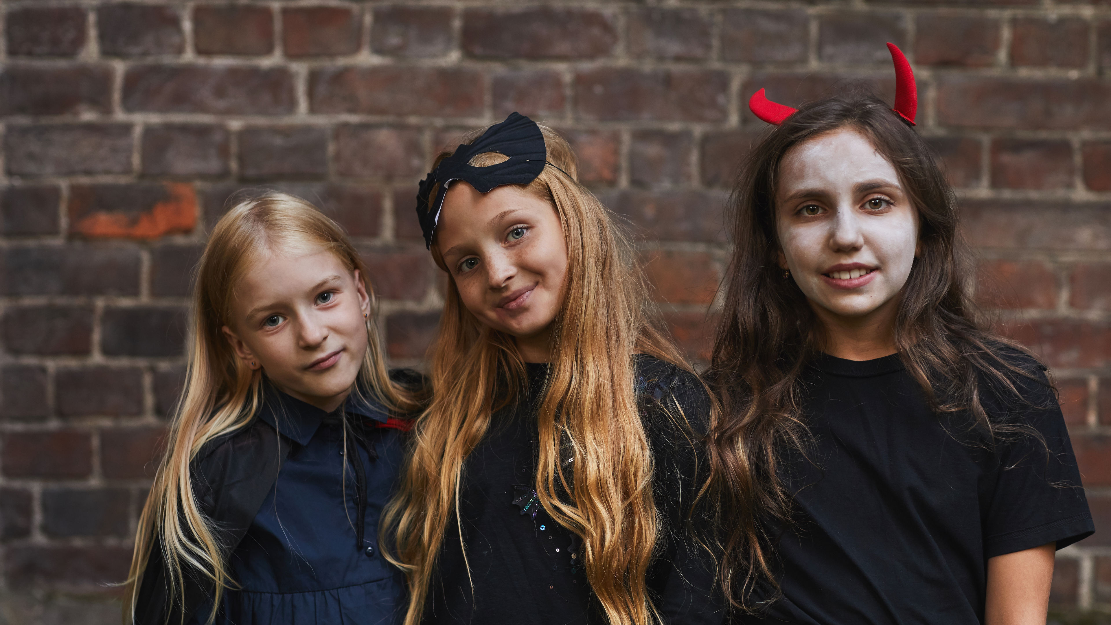 Three girls dressed in Halloween costumes, with one as a devil, another in a mask, and the third in a black cape 