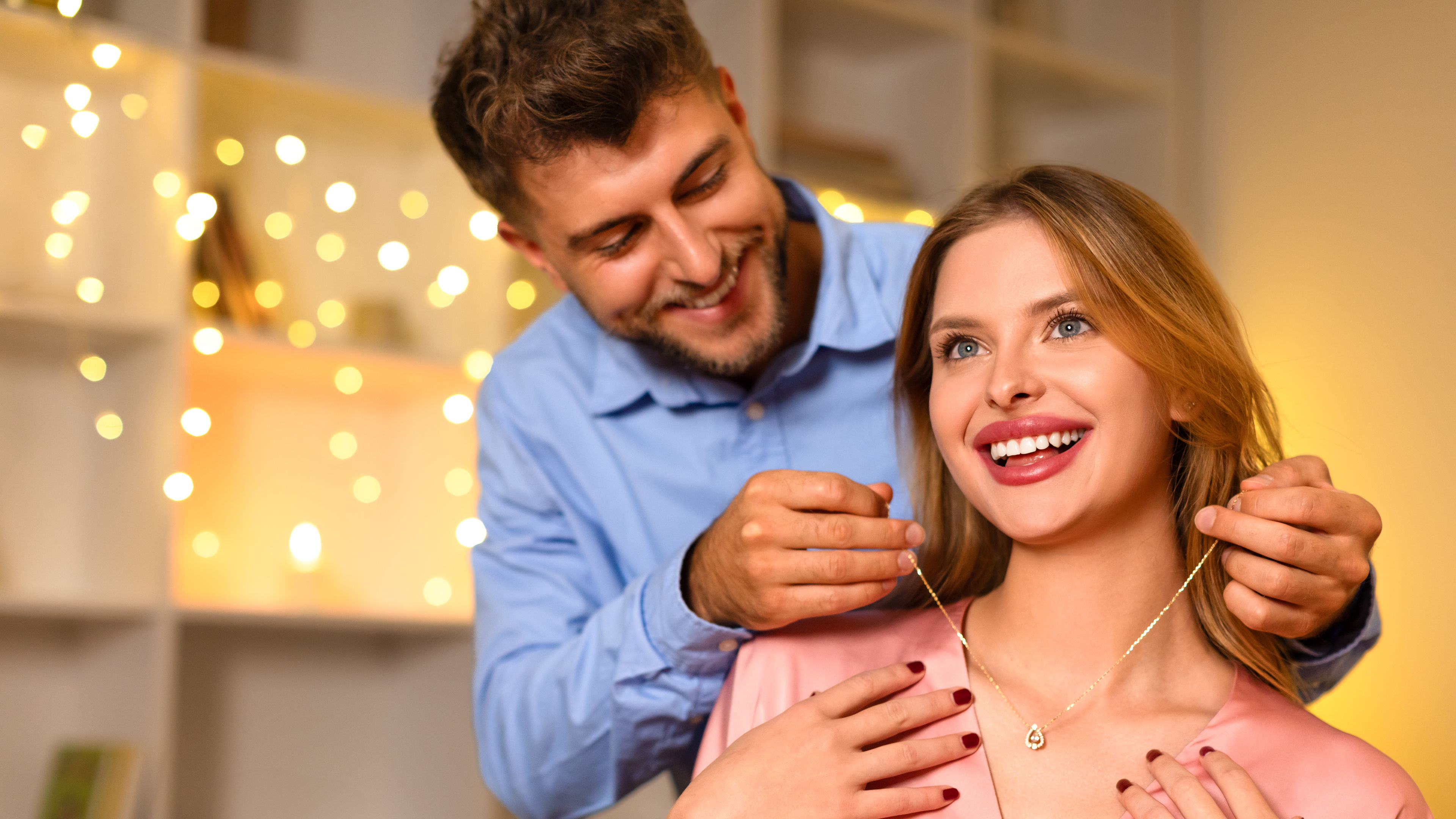 A smiling man places a gold necklace around a delighted woman’s neck  