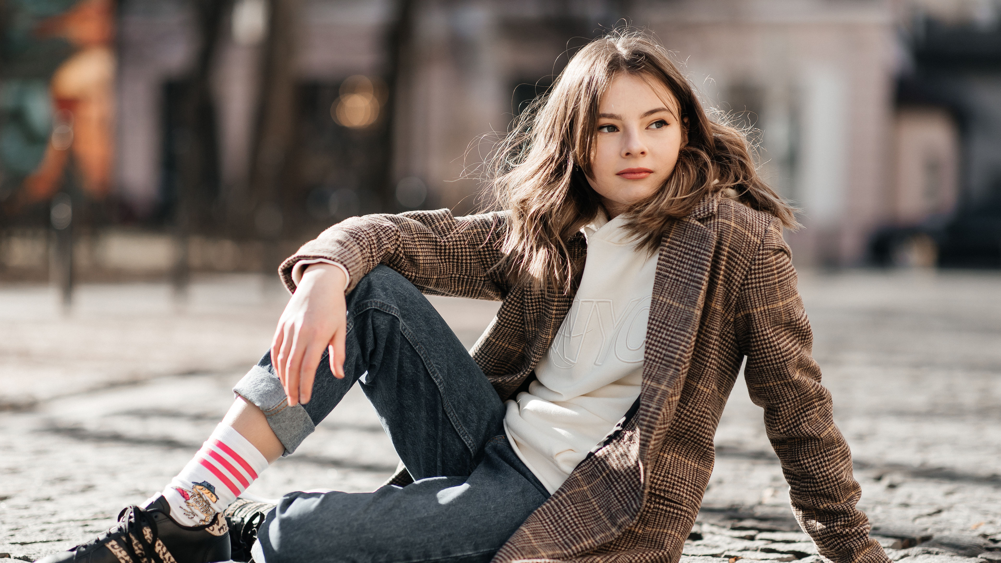 Stylish young woman sitting on a cobblestone street, effortlessly blending casual and chic with a plaid coat, hoodie, and trendy sneakers. 