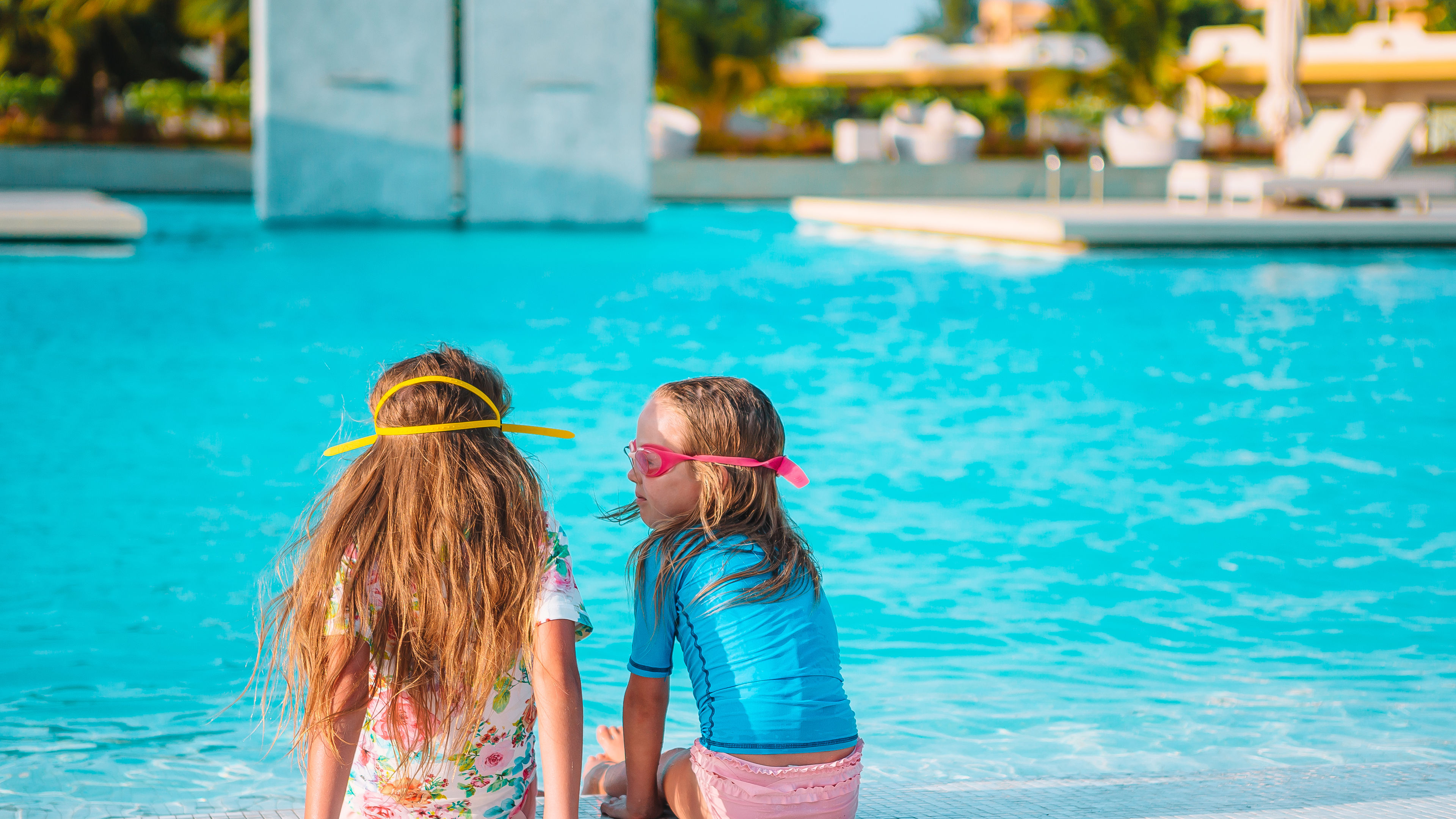 Two young girls in swimwear sit at the edge of a pool, chatting and enjoying the sunny day 