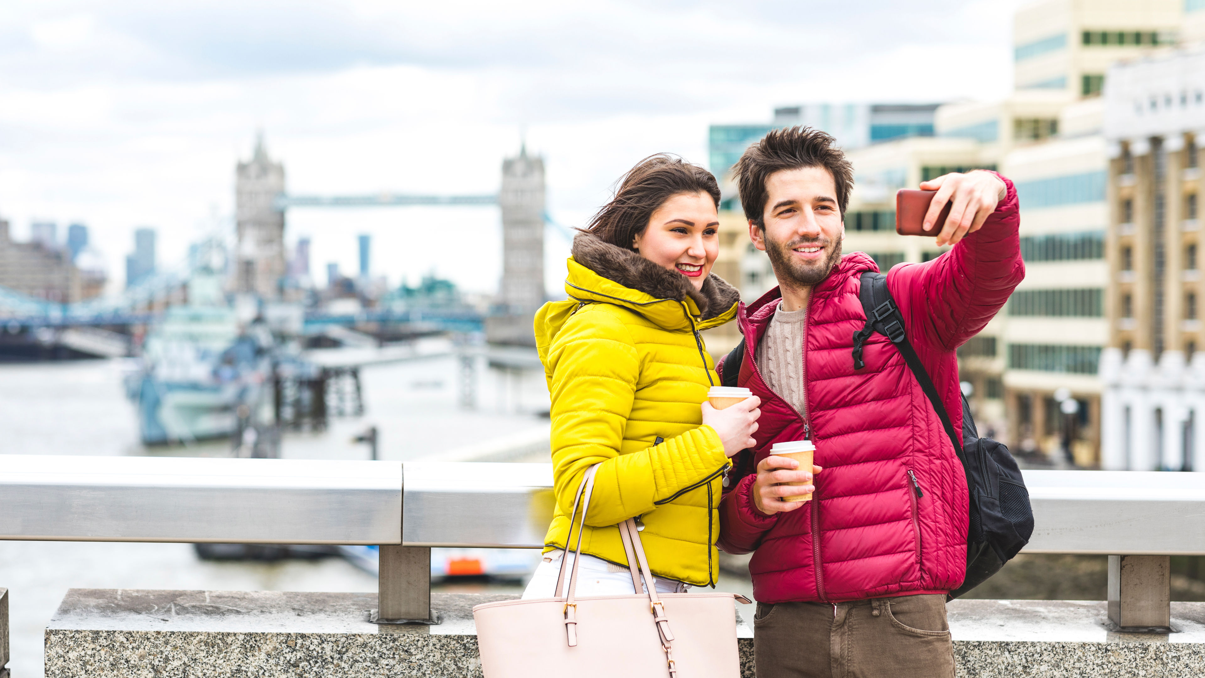 UK, London, young couple with coffee to go standing on bridge over the Thames taking selfie with smart phone