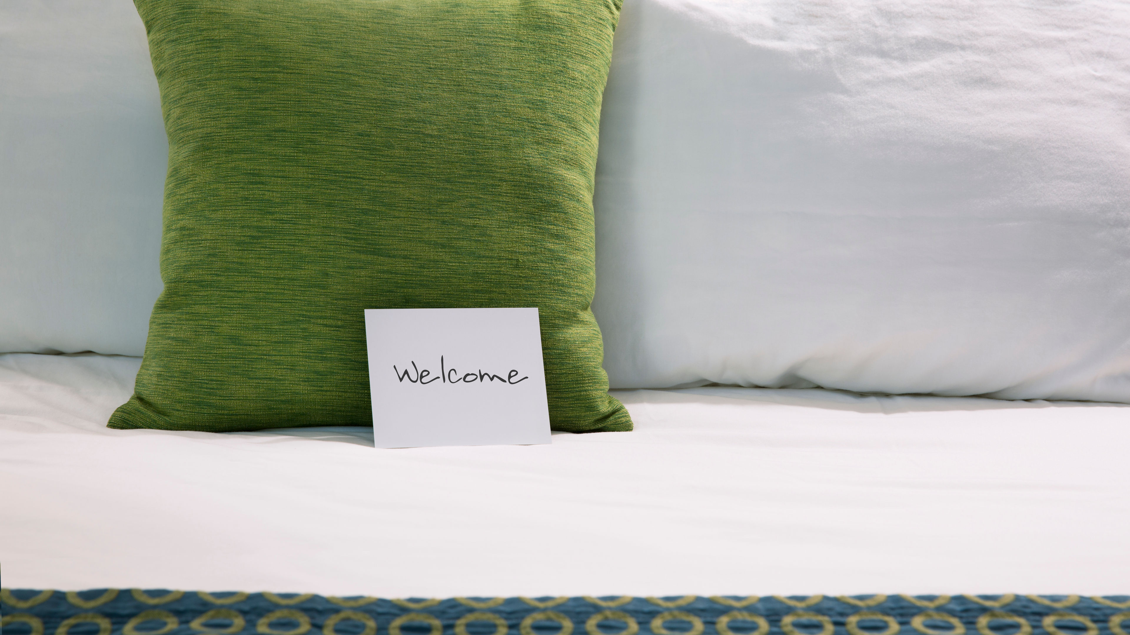 A green pillow and a "Welcome" card are placed neatly on a freshly made hotel bed 