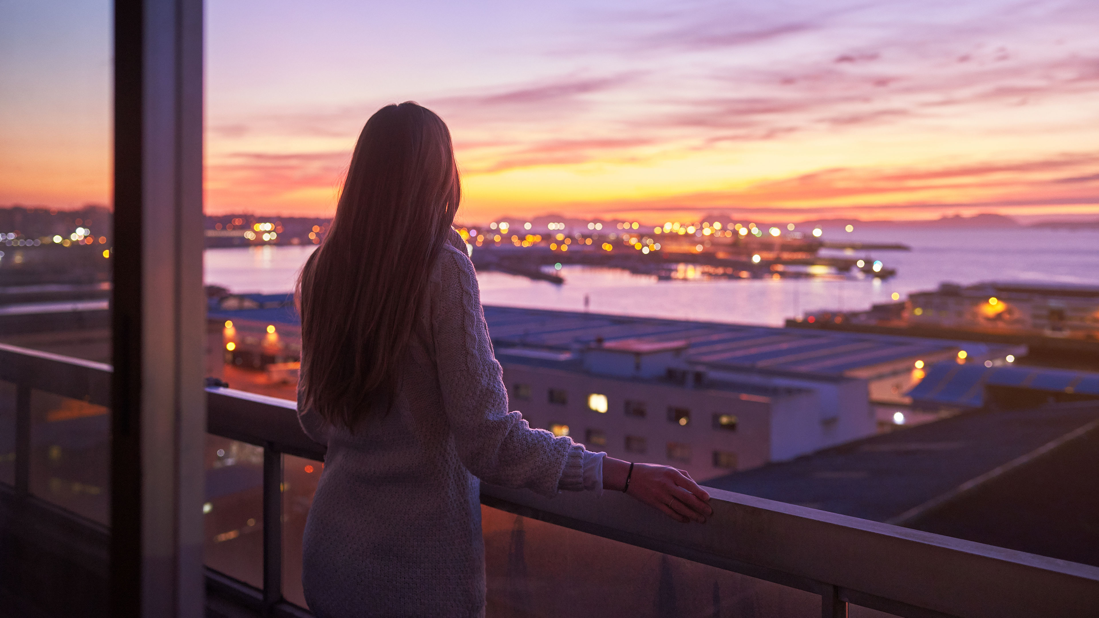 A woman leans on a balcony railing, watching a colorful sunset 