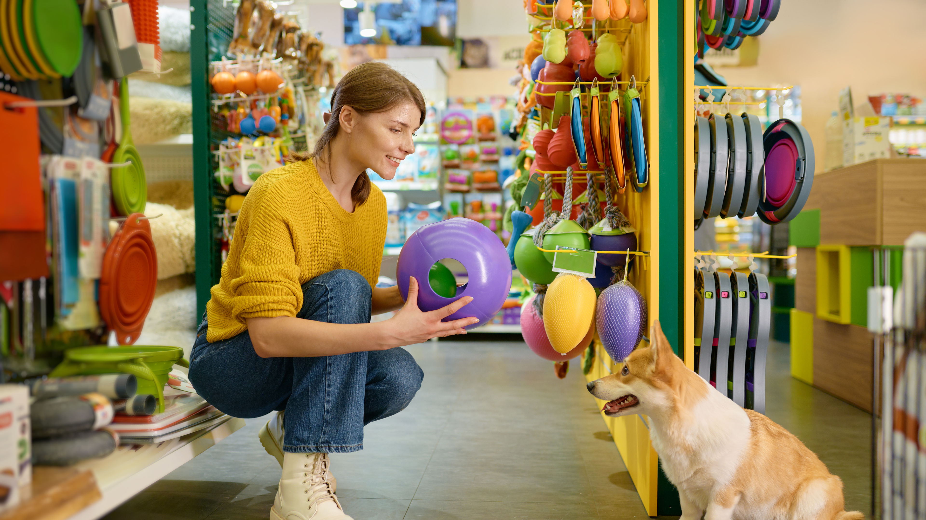 A woman in a yellow sweater crouches in a vibrant pet store aisle, selecting a purple toy ball for her happy pet dog 