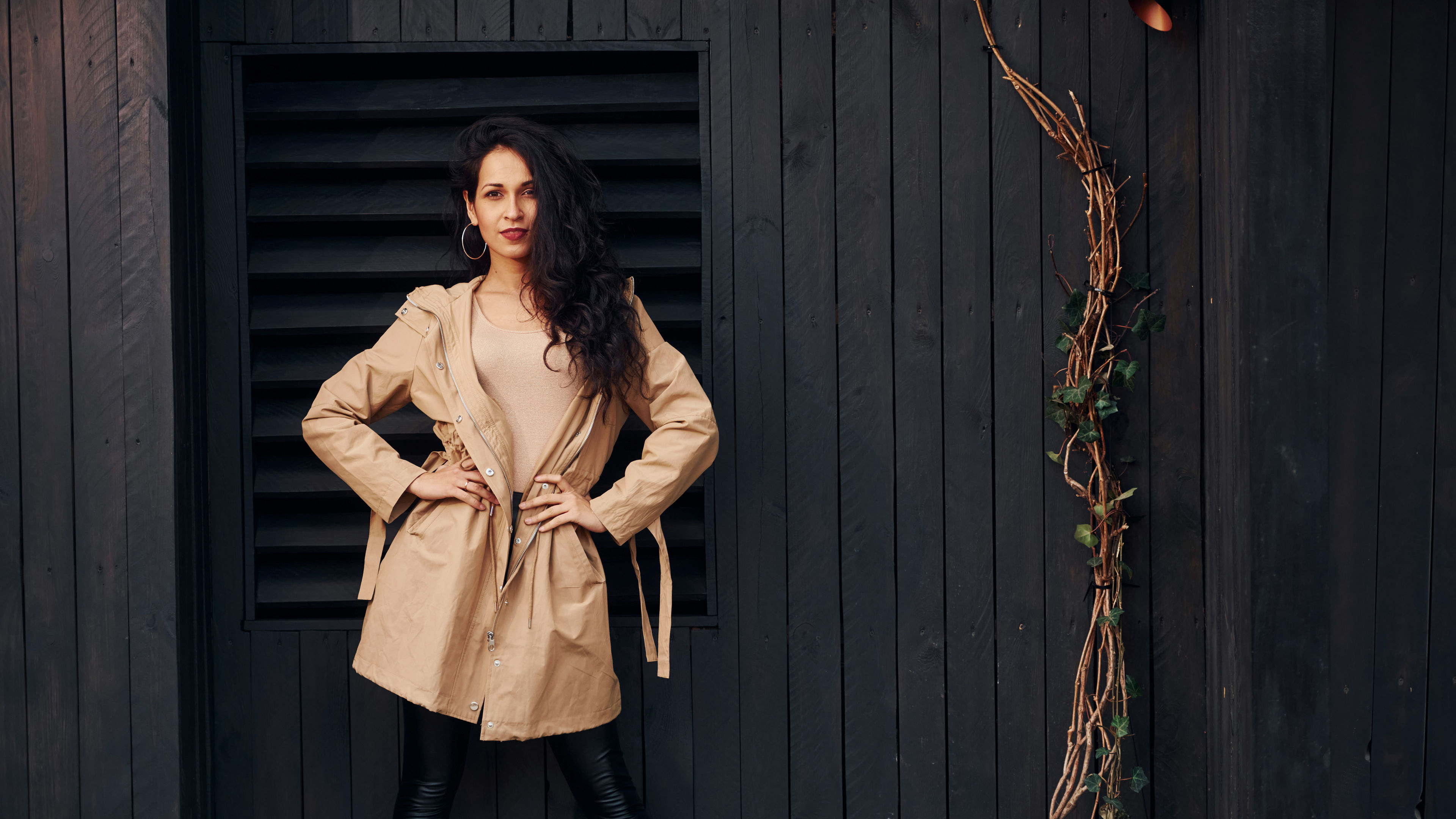 A woman with black curly hair stands against a black wooden building, her eyes reflecting the evening light. 